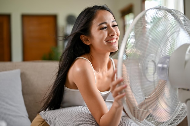 Una felice donna asiatica respira aria fresca da un ventilatore elettrico mentre riposa su un divano