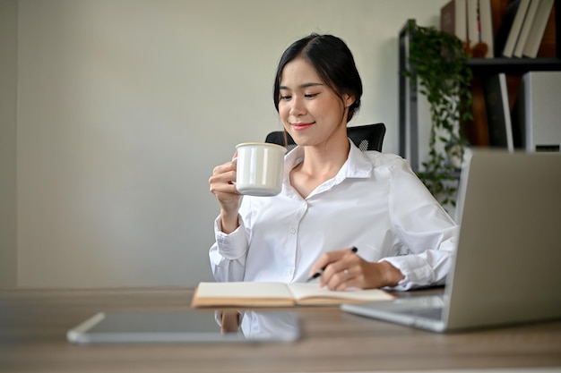 Una felice donna d'affari asiatica che si gode il caffè mattutino alla sua scrivania nel suo ufficio