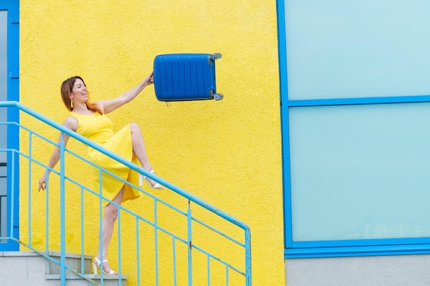 Una felice donna dai capelli rossi con un vestito giallo sta scendendo le scale e fa oscillare con gioia una valigia blu La ragazza si sta preparando per il viaggio Concetto di vacanza estiva
