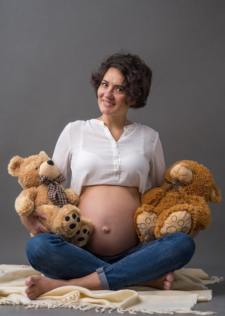 Una felice donna incinta sorridente si siede con la pancia esposta tenendo in mano degli orsacchiotti