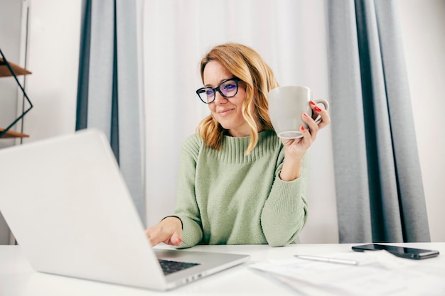 Una felice imprenditrice che lavora online sul suo laptop dall'home office e beve caffè