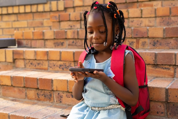 Una felice studentessa afroamericana seduta sulle scale e che usa lo smartphone a scuola elementare.