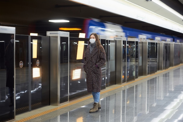 Una foto a figura intera di una donna con una maschera medica in attesa di un treno in arrivo sul binario della metropolitana