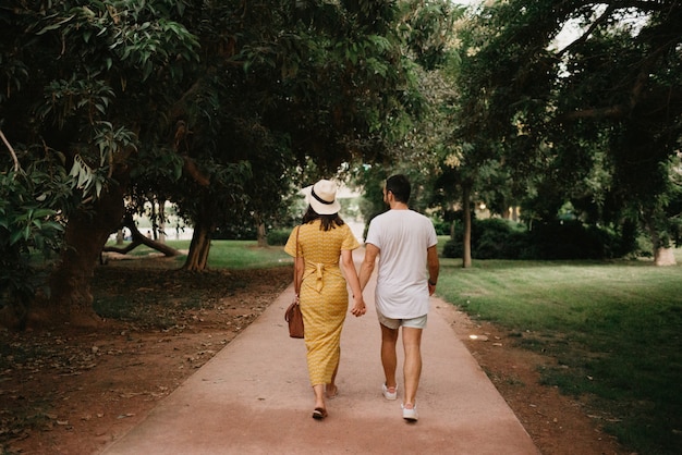 Una foto da dietro di una ragazza bruna con un vestito giallo e il suo ragazzo che camminano su un sentiero di sabbia tra gli alberi nel parco di Valencia. Un paio di turisti in una data in estate la sera.