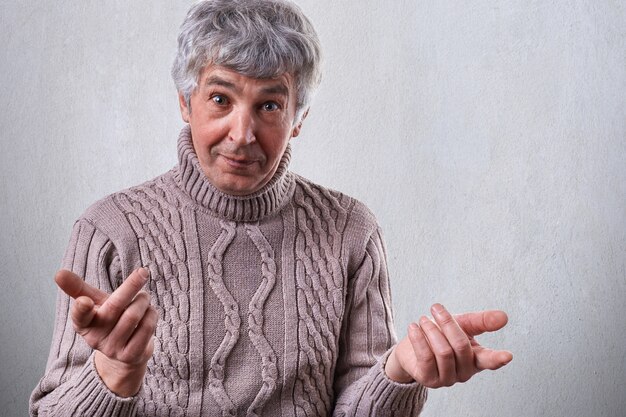 Una foto di un uomo maturo vestito di maglione in piedi sul muro bianco con espressione sorpresa tenendo le mani davanti a lui