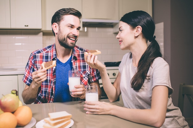 Una foto di una bella coppia si siede al tavolo e si guarda. Tengono toast con pasta di cioccolato su di esso. Inoltre ci sono bicchieri di latte nelle loro mani. Le persone sembrano felici.