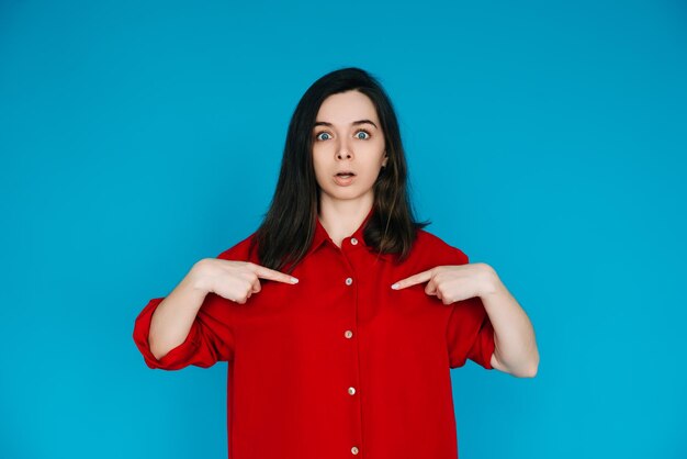 Una foto di una bella giovane donna colpita in una camicia rossa faccia sorpresa che punta il dito isolato su uno sfondo blu perfetto per enfatizzare sorpresa e stupore