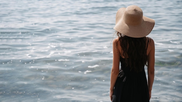 Una foto di una bella giovane donna con lunghi capelli scuri in un vestito e un cappello di paglia in posa sulla spiaggia in piedi con la schiena e guardando il mare infinito Un posto per il tuo testo