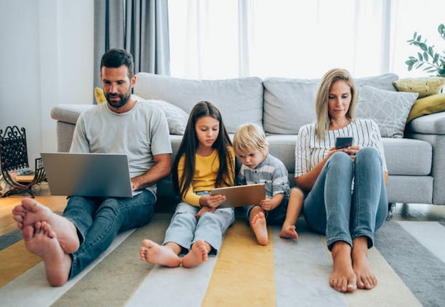 Una foto di una giovane famiglia sorridente con due bambini seduti sul pavimento nel soggiorno e che usano diversi dispositivi