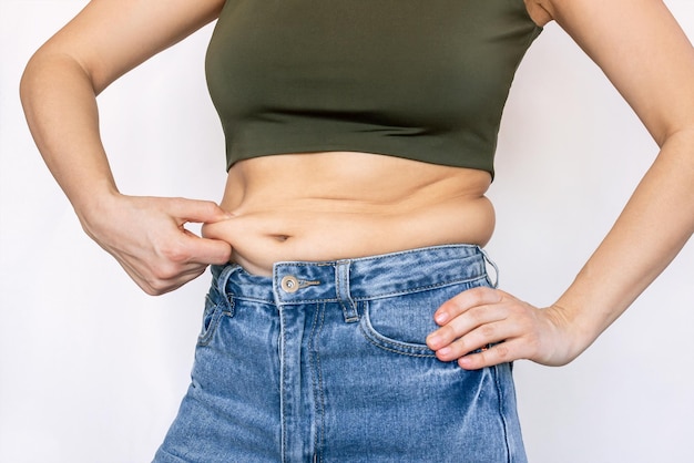 Una foto ritagliata di una giovane donna con un top corto e jeans che si tiene per il grasso sulla pancia