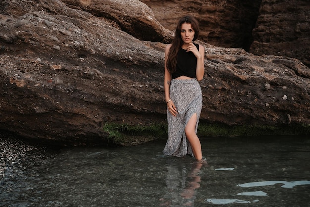 Una foto sulla spiaggia di una bella giovane donna sensuale con lunghi capelli scuri in un vestito che posa sulla spiaggia vicino a grandi massi