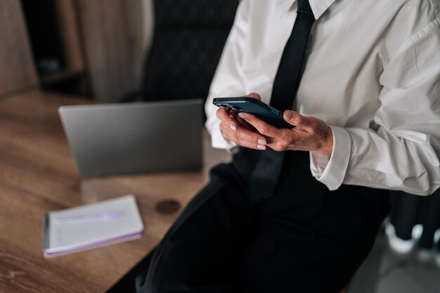 Una foto tagliata in primo piano di una donna d'affari irriconoscibile seduta su un tavolo d'ufficio che guarda il cellulare