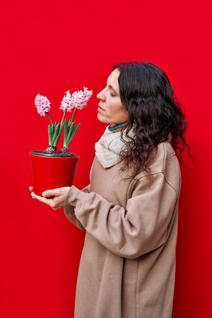 Una foto verticale di una bella donna odora un vaso con piante di giacinto sulla parete rossa