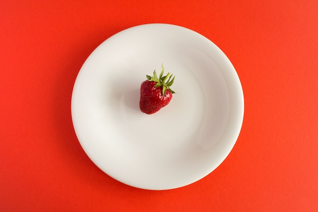 Una fragola nel piatto bianco sulla superficie rossa Vista dall'alto. Concetto minimo diabolico.