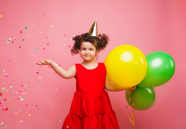 Una gioiosa bambina con un vestito rosso festeggia il suo compleanno e fa uscire coriandoli colorati su uno sfondo rosa un bambino tiene palloncini e cattura coriandoli