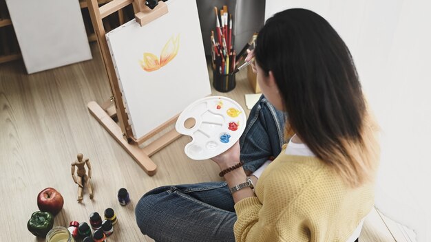 Una giovane artista femminile con una tavolozza in mano che dipinge su tela mentre è seduta sul pavimento nel suo laboratorio.