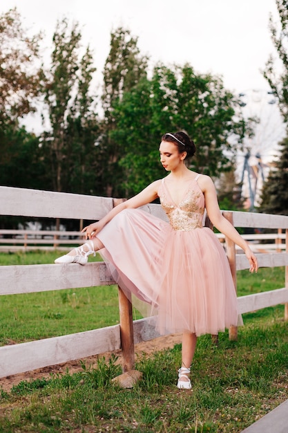 Una giovane ballerina elegante mette il piede su una staccionata di legno e annoda i nastri delle scarpe da punta in pa...