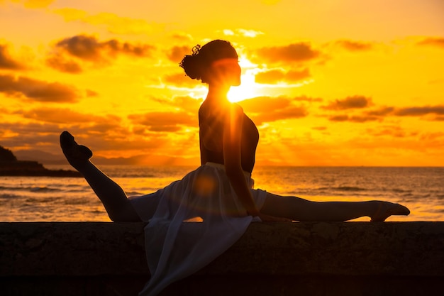 Una giovane ballerina sulla spiaggia al tramonto che fa una spagata con il mare sullo sfondo