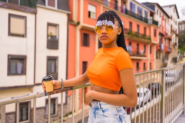 Una giovane ballerina trap con le trecce. Ragazza etnica nera con t-shirt, occhiali da sole arancioni e pantaloni da cowboy con tazza Take-Away in città