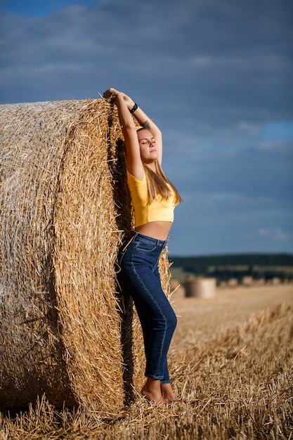 Una giovane bella bionda si erge su un campo di grano falciato vicino a un enorme covone di fieno, godendosi la natura. Natura nel villaggio