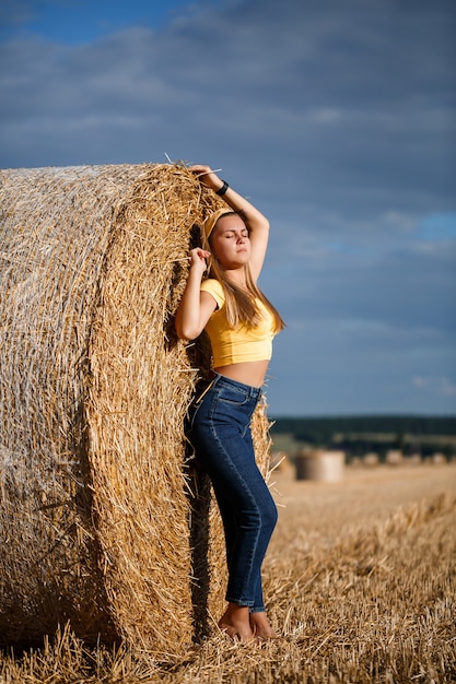 Una giovane bella bionda si erge su un campo di grano falciato vicino a un enorme covone di fieno, godendosi la natura. Natura nel villaggio