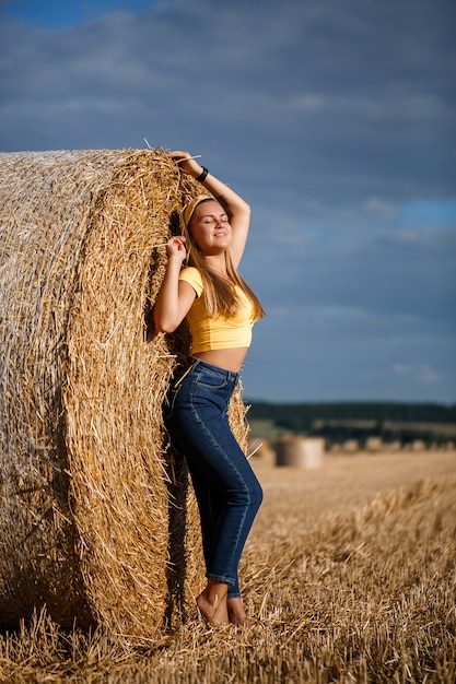 Una giovane bella bionda si erge su un campo di grano falciato vicino a un enorme covone di fieno, godendosi la natura. Natura nel villaggio