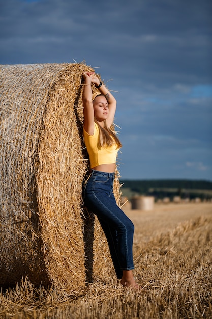 Una giovane bella bionda si erge su un campo di grano falciato vicino a un enorme covone di fieno, godendosi la natura. Natura nel villaggio