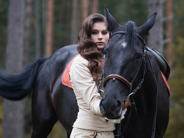 Una giovane bella bruna cavaliere accanto a un cavallo nero in pieno di munizioni ritratto in una stalla in un ...