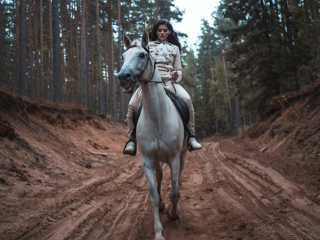 Una giovane bella bruna cavaliere in un elegante abito retrò a cavallo bianco in un'area forestale