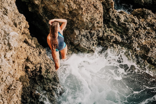 Una giovane bella donna abbronzata con i capelli lunghi in bikini blu seduta sulle rocce vicino al verde blu chiaro