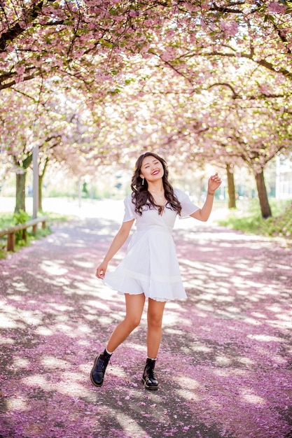 Una giovane bella donna asiatica in un abito bianco cammina in un parco fiorito. Sakura. Alberi in fiore. Primavera.