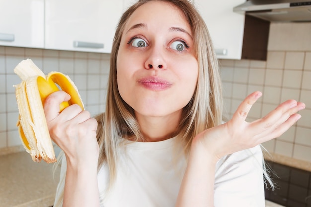 Una giovane bella donna bionda caucasica in una maglietta bianca mangia una banana e ne gode il gusto in cucina