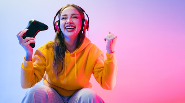 Una giovane bella donna caucasica con una felpa con cappuccio gialla che gioca a un videogioco utilizzando un joystick e cuffie con una divertente sorpresa di messa a fuoco e una faccia vittoriosa vincente con luce al neon blu e rossa