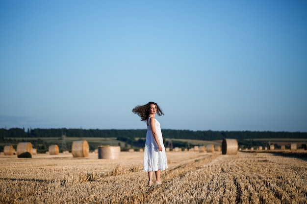 Una giovane bella donna in un abito estivo bianco si trova su un campo di grano falciato con enormi covoni di fieno, godendosi la natura. Natura nel villaggio. Messa a fuoco selettiva