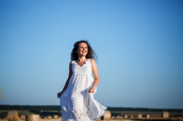 Una giovane bella donna in un abito estivo bianco si trova su un campo di grano falciato con enormi covoni di fieno, godendosi la natura. Natura nel villaggio. Messa a fuoco selettiva