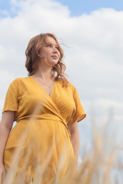Una giovane bella donna incinta in un vestito giallo e un cappello cammina attraverso un campo di grano arancione in una soleggiata giornata estiva
