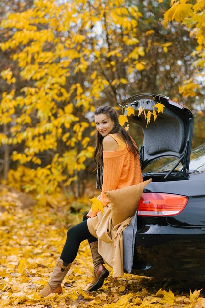 Una giovane bella donna si siede sul bagagliaio di un'auto nella foresta d'autunno