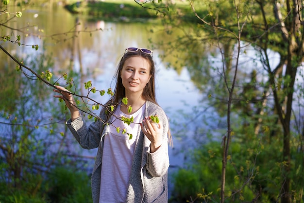 Una giovane bella ragazza con gli occhiali da sole sta camminando vicino al lago, con in mano un ramo di un albero