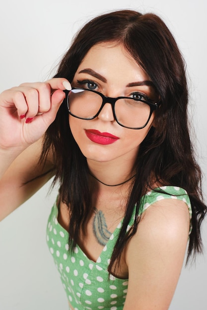 Una giovane bella ragazza con gli occhiali guarda accigliata la telecamera su uno sfondo chiaro in un abito a pois