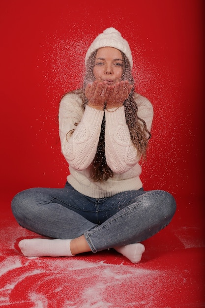 una giovane bella ragazza con un cappello invernale bianco e un maglione lavorato a maglia soffia sulla neve artificiale