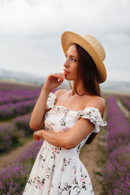 Una giovane bella ragazza con un vestito e un cappello delicati cammina attraverso un bellissimo campo di lavanda e gode dell'aroma dei fiori. Vacanza e natura meravigliosa.