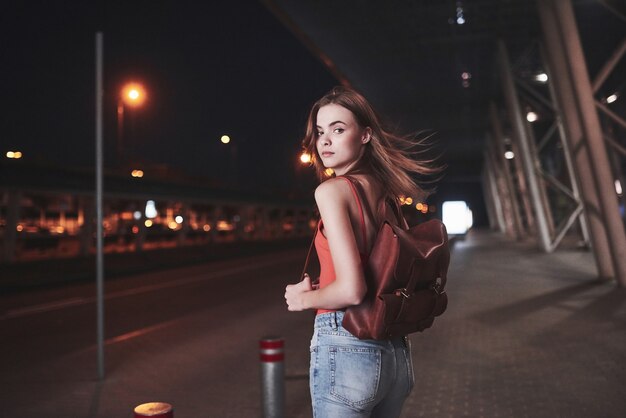 Una giovane bella ragazza con uno zaino dietro le spalle si trova per strada vicino a un aeroporto o una stazione ferroviaria in una calda sera d'estate. È appena arrivata e aspetta un taxi o i suoi amici.