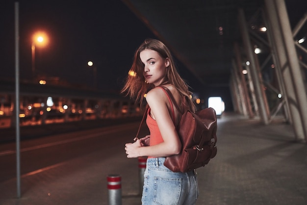 Una giovane bella ragazza con uno zaino dietro le spalle si trova per strada vicino a un aeroporto o una stazione ferroviaria in una calda sera d'estate. È appena arrivata e aspetta un taxi o i suoi amici.