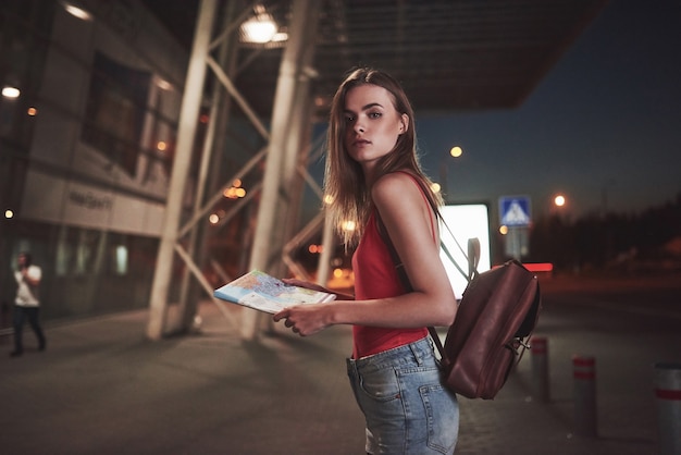 Una giovane bella ragazza con uno zaino dietro le spalle si trova per strada vicino a un aeroporto o una stazione ferroviaria in una calda sera d'estate. È appena arrivata e aspetta un taxi o i suoi amici.
