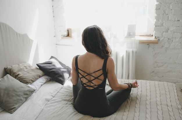 Una giovane bella ragazza in abiti sportivi fa yoga a casa in autoisolamento, un atleta medita in una stanza al mattino, vista posteriore