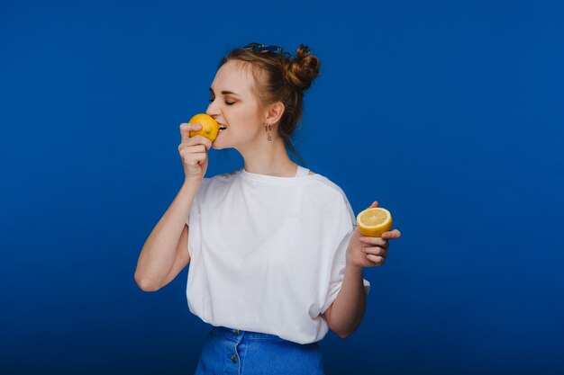 Una giovane bella ragazza in piedi su uno sfondo blu che tiene i limoni in mano e morde.