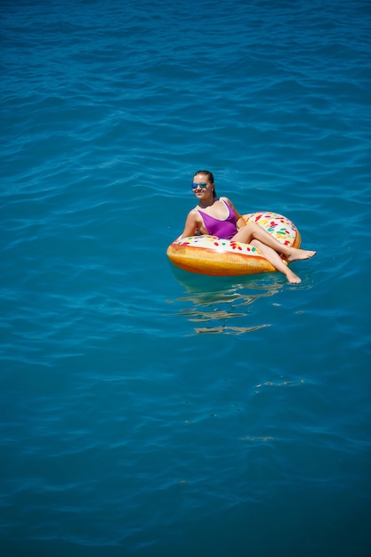 Una giovane bella ragazza in un costume da bagno luminoso giace su un grande anello gonfiabile e galleggia sul mare blu in una luminosa giornata estiva di sole