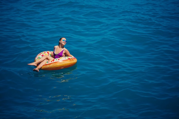 Una giovane bella ragazza in un costume da bagno luminoso giace su un grande anello gonfiabile e galleggia sul mare blu in una luminosa giornata estiva di sole