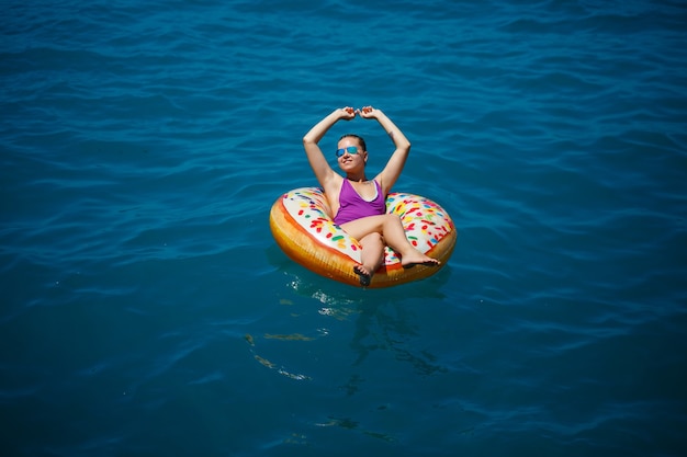 Una giovane bella ragazza in un costume da bagno luminoso si trova su un grande anello gonfiabile e galleggia sul mare blu in una luminosa giornata estiva di sole. Messa a fuoco selettiva