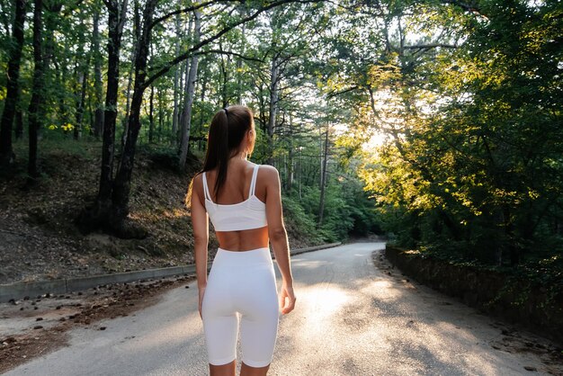 Una giovane bella ragazza posa con la schiena prima di correre l'allenamento su strada in una fitta foresta durante il tramonto Uno stile di vita sano e correre all'aria aperta